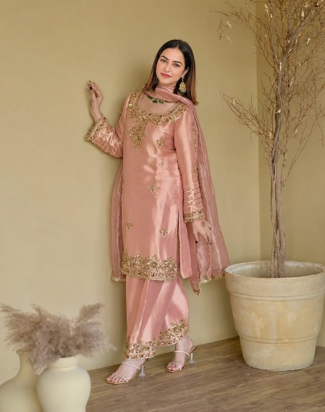 Peach-pink metallic kurta set with gold hand embroidery and a matching dupatta, worn against an ochre wall beside a dried-branch plant