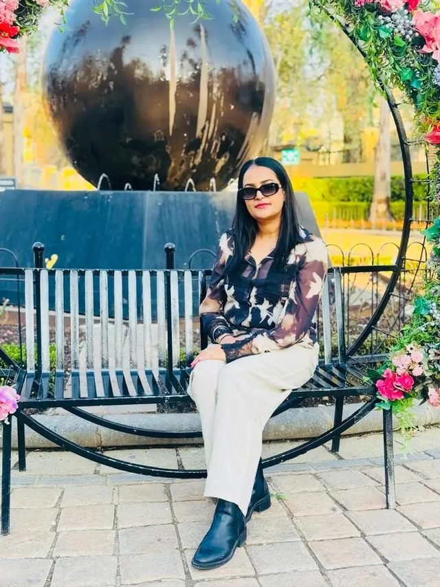 Woman in sunglasses sitting on a garden bench wearing a sheer printed shirt and cream trousers, framed by flowers